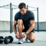 Athletic man tying shoelaces next to dumbbells in a minimalist outdoor workout area, with a blurred pull-up bar in the background.